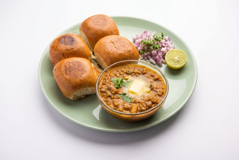 Pav Bhaji, a popular Indian street food, with butter, chopped onions, and lime on a green plate.